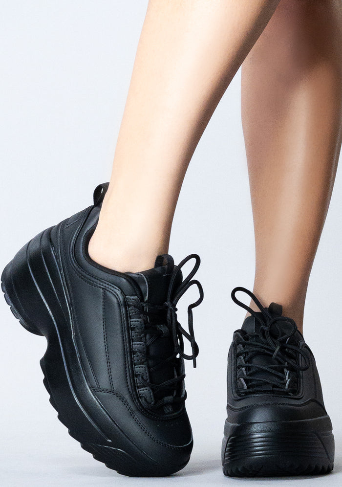 Front view of the black LILY 5005 Next Level platform sneakers on-model, showing the clean tongue design, black laces, and wide toe box.