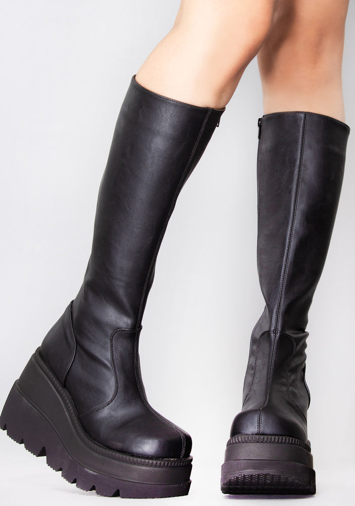 Demonia SHAKER 100 Total Blackout Black Platform Boots in black vegan PU leather with stacked wedge platform worn by model — front view