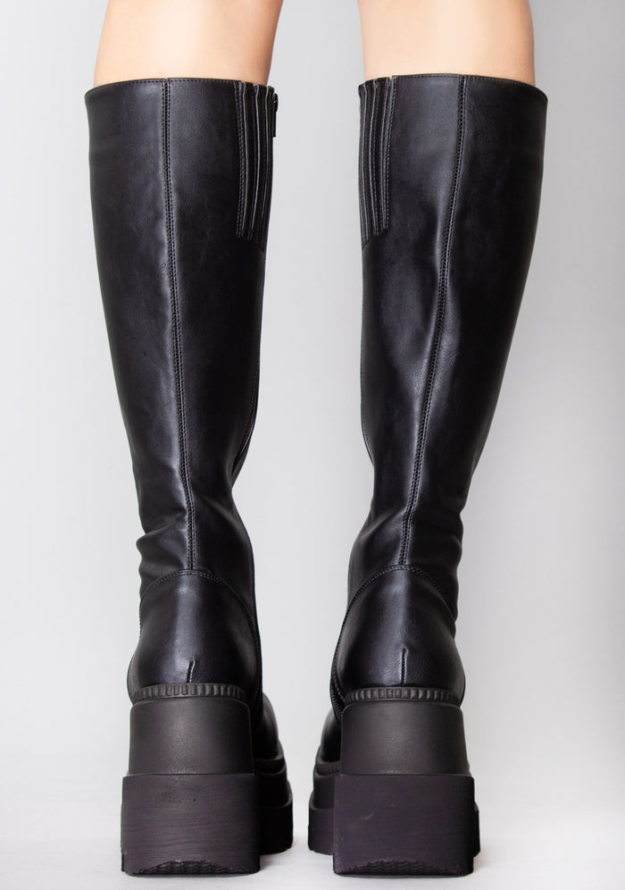Demonia SHAKER 100 Total Blackout Black Platform Boots in black vegan PU leather with knee-high silhouette worn by model — back view